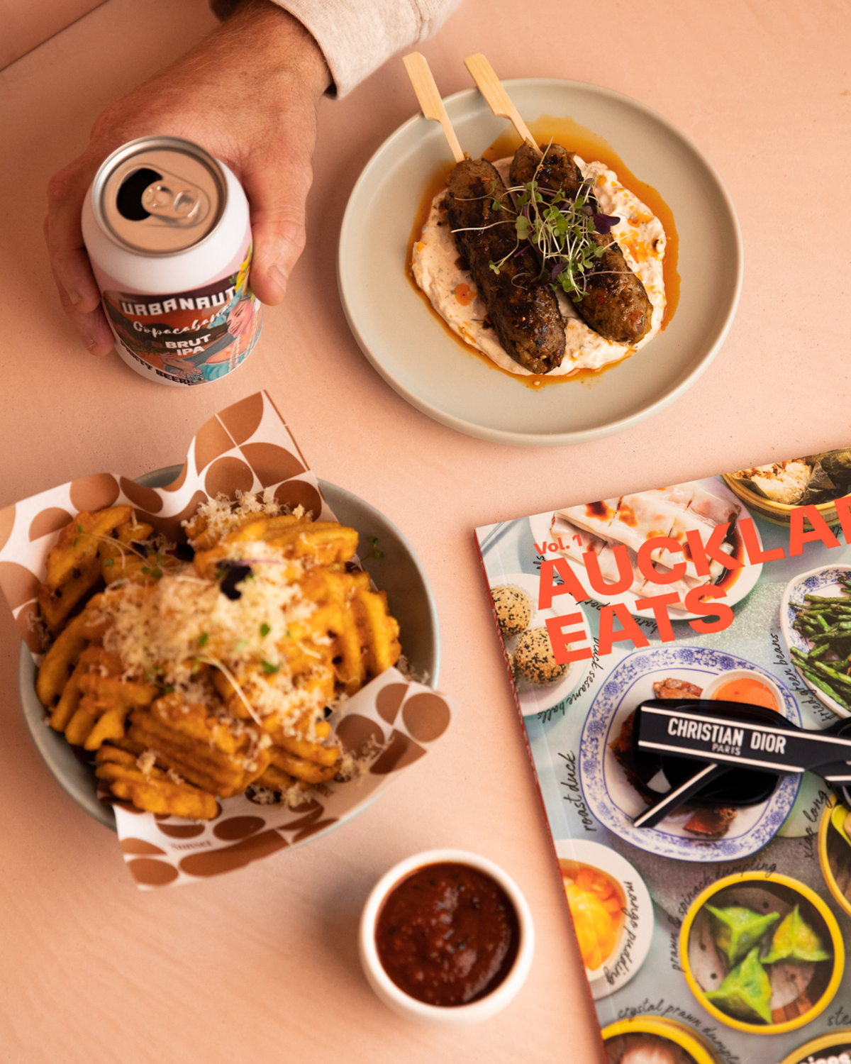 Stylish aerial view of hand holding beer in top left, with bowl of waffle fries with parmesan cheese topping, lamb skewers on hummus and Auckland Eats magazine with Christian Dior glasses on top 